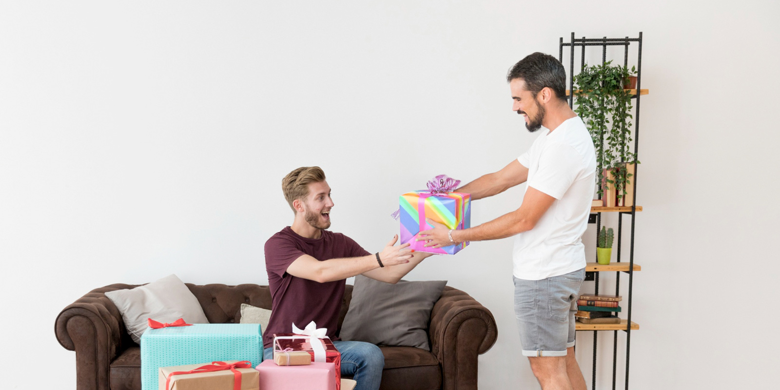 Homem sorridente entregando um presente com embrulho de arco-íris a outro homem sentado em um sofá, representando um momento de amizade e celebração.