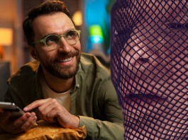 Homem sorrindo enquanto navega no celular, com uma mulher em meia arrastão ao fundo, simbolizando o ato de espionar perfis em plataformas como OnlyFans.