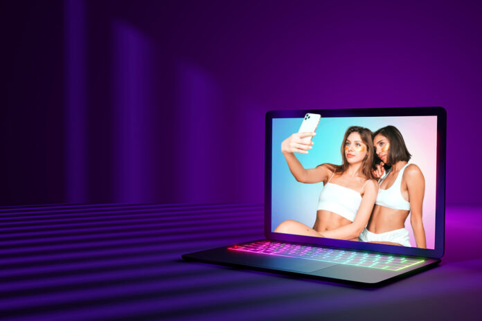 Duas mulheres jovens tirando selfie exibidas na tela de um notebook moderno com fundo roxo iluminado, representando interação digital e plataformas online.
