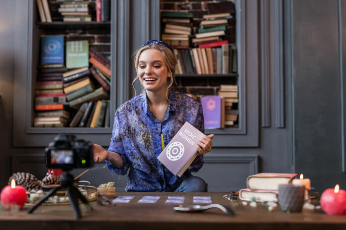 criadora-gravando-curso-online-em-2025 Mulher jovem sorridente gravando curso online com câmera profissional em ambiente aconchegante e decorado com livros, representando ensino digital em 2025.