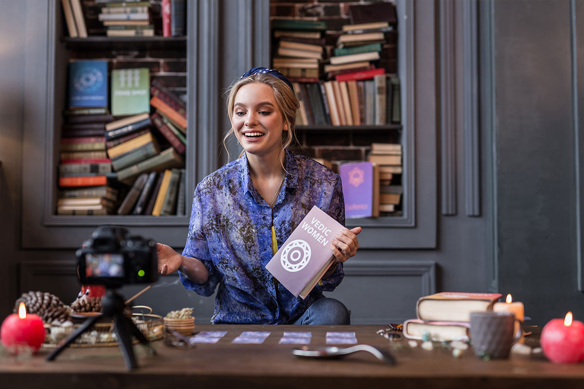 Mulher jovem sorridente gravando curso online com câmera profissional em ambiente aconchegante e decorado com livros, representando ensino digital em 2025.