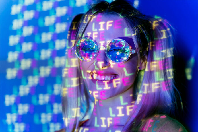criadora-conteudo-adulto-inteligencia-artificial Mulher sorridente com óculos futuristas refletindo a palavra "LIFE" em projeções coloridas, representando criatividade e uso de inteligência artificial no conteúdo digital.