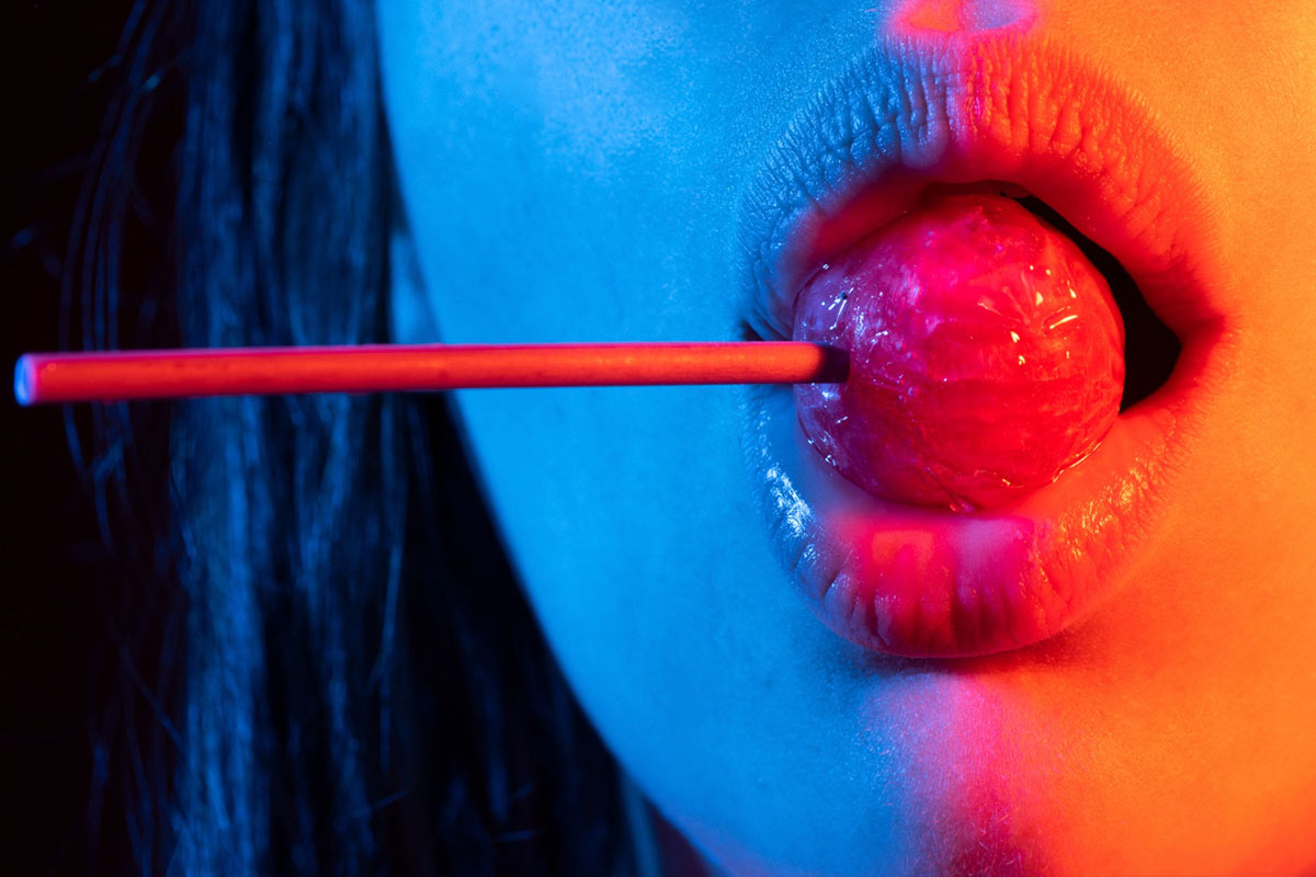 Close-up de lábios femininos segurando um pirulito em iluminação neon azul e vermelha, simbolizando sensualidade e estilo provocativo