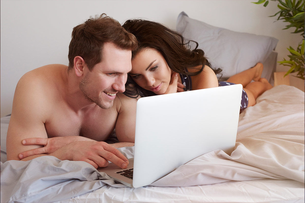 Casal deitado na cama usando notebook para explorar experiências e conexões online.