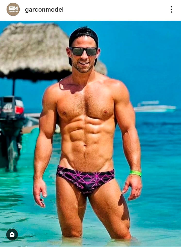 Homem usando sunga estampada da marca Garçon Model no mar, representando moda praia gay internacional em 2026